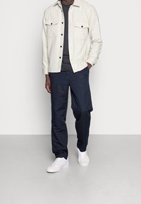 Hombre con una camisa de botón beige, camiseta gris oscuro, pantalones azul marino y zapatillas blancas, de pie con una mano en el bolsillo.