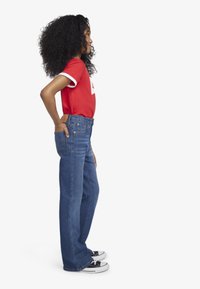 T-shirt rouge avec des accents blancs, jean en denim bleu à coupe droite et baskets noires avec semelles blanches. Vue de côté.