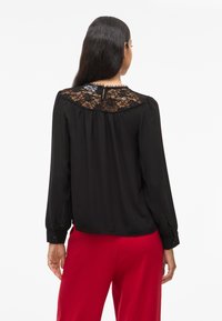 Blusa preta com detalhes em renda nos ombros, mangas longas, punhos com botões e um fechamento nas costas em forma de gota. Textura suave.