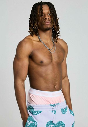 Mężczyzna bez koszulki z włosami do ramion, skręconymi, ubrany w warstwowe różowo-turkusowe spodenki w wzory, stojący z rękami w kieszeniach na szarym tle.