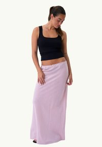 Γυναίκα που φορά μαύρο αμάνικο τοπ και μακριά ανοιχτό ροζ φούστα, στέκεται και κοιτάζει προς τα δεξιά και κάτω.