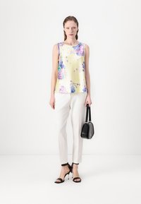 Top sin mangas floral en amarillo claro, con patrones florales en púrpura y verde. Combinado con pantalones blancos a medida y un bolso negro.