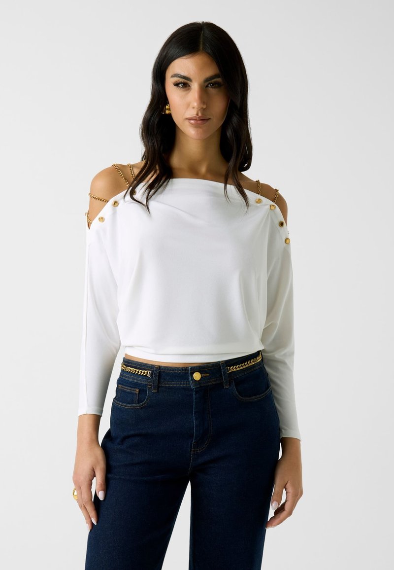 Femme portant un haut blanc à épaules dénudées avec des détails en boutons dorés et chaînes, associé à un jean bleu foncé avec une ceinture chaîne dorée.