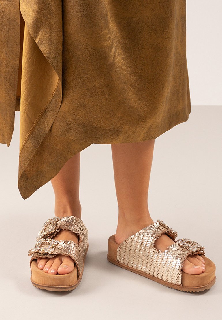 Sandalias deslizantes metálicas doradas tejidas con una suela gruesa. Cuentan con dos correas anchas adornadas con nudos decorativos. Acabado texturizado.
