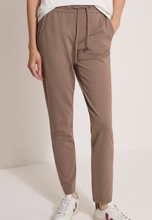 Personne portant un pantalon taupe fuselé avec taille à cordon et des baskets blanches avec des accents bordeaux, debout les mains dans les poches.