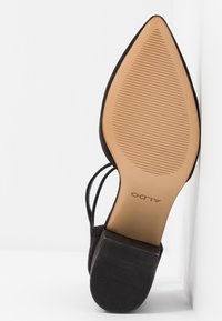 Semelle de chaussure noire à bout pointu avec un dessous beige texturé, talon carré, et deux fines brides visibles par en dessous.