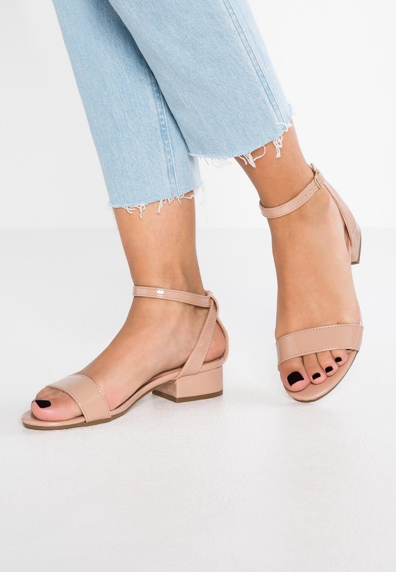 Sandales à talon bloc en cuir verni nude avec bride à la cheville, design à bout ouvert, et texture lisse. Portées avec un jean bleu clair effiloché.