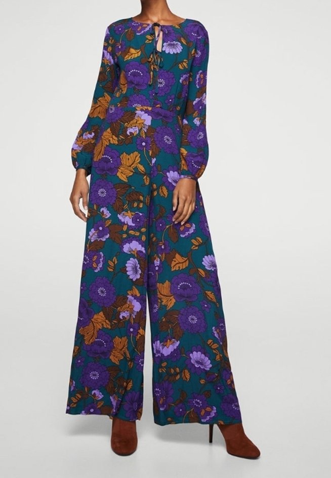 Femme portant une combinaison bleu sarcelle avec un grand imprimé floral violet et orange, manches longues et jambes larges, associée à des bottes marron à talons.