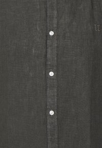 Camisa de lino gris oscuro con cierre de botones blancos en la parte delantera y detalles sutiles bordados en el pecho. Tejido texturizado.