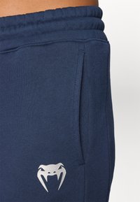 Tamno plave trenirke od mekog materijala, s bočnim džepovima i srebrnim logotipom blizu struka. Elastična waistband za sigurno prianjanje.