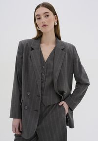 Blazer grigio a righe con rever a punta e chiusura a doppio bottone, abbinato a un gilet coordinato con scollo a V e dettagli con bottoni.
