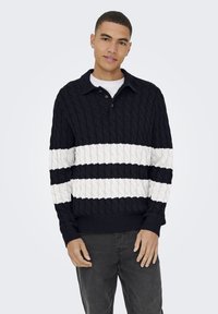 Suéter de punto cableado azul marino con rayas horizontales blancas y un cuello con solapa. Presenta puños y dobladillo acanalados, y una tapeta de dos botones.