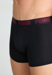 Boxeri negri dintr-un material neted, cu un elastic lat de culoare maroon pe care este imprimat "PUMA" cu litere îndrăznețe, în contrast.