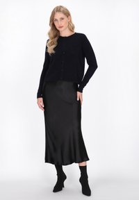 Marineblauwe button-up cardigan met een ronde hals, gecombineerd met een zwarte satijnen midi rok en zwarte strakke soklaarzen. De rok heeft een gladde textuur.