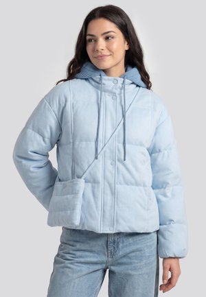 Young woman wearing a light blue puffer jacket with a matching crossbody bag and blue jeans, looking to the side with a slight smile.