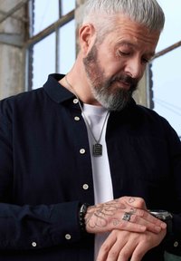 Black button-up shirt over a white T-shirt, featuring silver jewelry, a pendant necklace, and a textured wristband on a tattooed wrist.