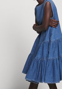 Džínové šaty s víceúrovňovým designem, bez rukávů a kulatým výstřihem. Světle modrá barva s jemnými stehy a rozšířenou sukní.