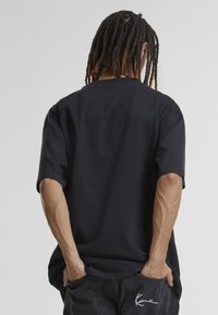 T-shirt noir oversize à manches courtes, en coton, col rond, avec un détail de couture subtil au dos. Porté avec un pantalon foncé.