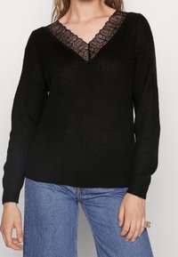 Femme portant un pull en tricot noir avec un détail en dentelle en V et un jean bleu, montrant le haut du torse et la main droite avec une bague.