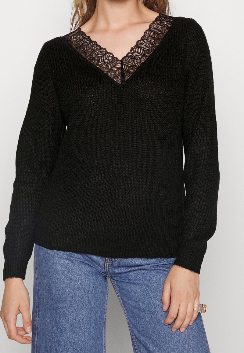 Femme portant un pull en tricot noir avec un détail en dentelle en V et un jean bleu, montrant le haut du torse et la main droite avec une bague.