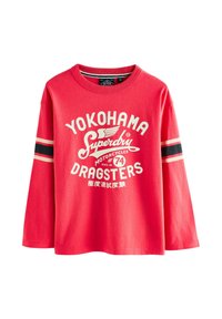 Rød bluse med lange ærmer i bomuld. Har et hvidt grafisk tryk med teksten "YOKOHAMA SUPERDRY DRAGSTERS" og sorte vandrette striber på ærmerne.