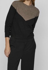 Donna che indossa un maglione color block nero e marrone con un design diagonale, abbinato a pantaloni neri su misura, su uno sfondo neutro.