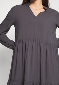 Femme portant une robe ample à manches longues, gris foncé, avec un col en V et un détail froncé à la taille, debout les mains sur les hanches.