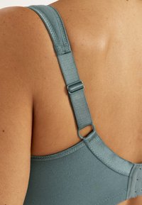 Makroaufnahme eines verstellbaren blauen BH-Trägers auf dem Rücken einer Person mit sichtbaren Nähten und Verschluss-Haken.