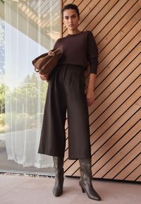 Brown knit sweater, gray pleated wide-leg trousers, green knee-high heeled boots, holding a brown suede bag, with wooden background.