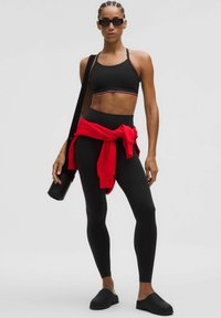Sujetador deportivo negro con acento rojo, leggings negros de talle alto, sudadera roja atada a la cintura y zapatos negros sin cordones.