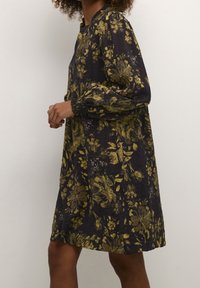 Mujer vistiendo un vestido negro con un patrón floral dorado y mangas largas abullonadas, de pie contra un fondo claro y liso.