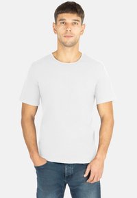 Junger Mann in schlichtem weißen T-Shirt mit kurzen Ärmeln und blauen Jeans steht vor einem hellen Hintergrund mit einer Hand in der Tasche.