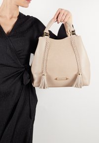 Borsa beige realizzata in materiale testurizzato, con dettagli intrecciati e frange, rifinita con bordo marrone e hardware dorato.