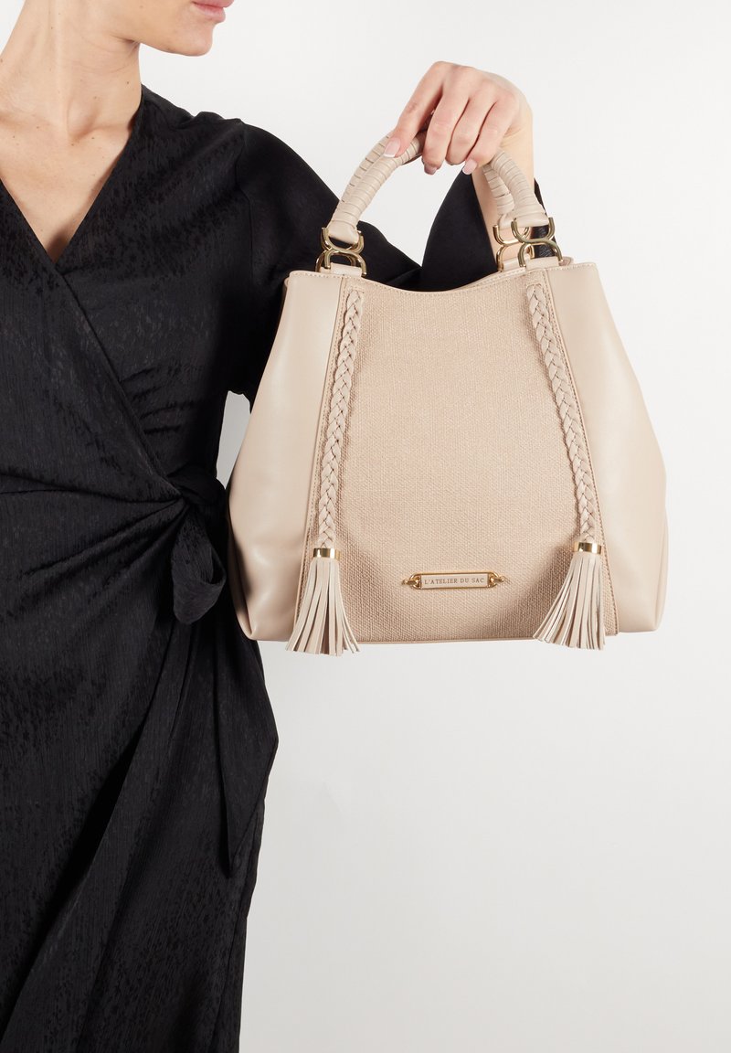 Borsa beige realizzata in materiale testurizzato, con dettagli intrecciati e frange, rifinita con bordo marrone e hardware dorato.