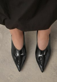 Zapatos de tacón puntiagudos de charol negro con un acabado suave y brillante y lados abiertos, posicionados sobre una superficie gris.