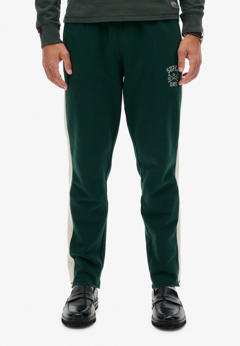 Grüne Jogginghose mit cremefarbenen Seitenstreifen und einem Logo auf dem linken Oberschenkel. Weicher Stoff, tapered Fit und elastischer Bund für zusätzlichen Komfort.