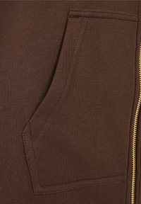 Poche de sweat-shirt en tissu marron avec coutures visibles et vue partielle d'une fermeture éclair dorée sur le côté droit.