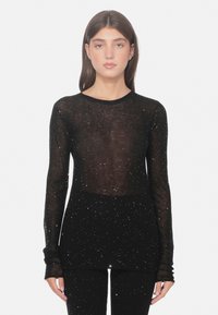 Vrouw in een zwarte transparante lange mouwtop en bijpassende broek met verspreide kleine witte glinsteringen, staand tegen een eenvoudige witte achtergrond.