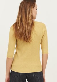 Gul ribbet strikbluse med trekvartærmer, struktureret stof, tætsiddende design og lige søm, iført med mørke jeans.