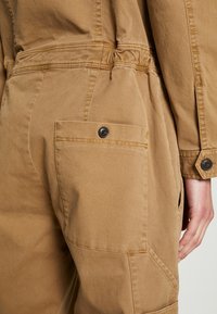 Primer plano de pantalones de trabajo color beige con un bolsillo con solapa asegurado por un botón negro, llevados por una persona con la mano relajada a un lado.