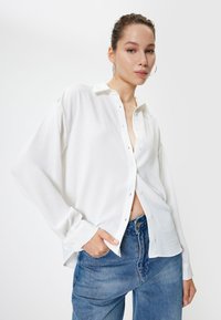 Weißes Button-Down-Hemd aus glattem Stoff, mit Kragen, langen Ärmeln und einer lockeren Passform, getragen mit blauen hoch taillierten Jeans.