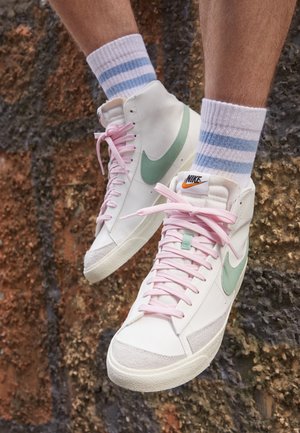 Stopy w białych wysokich tenisówkach Nike z różowymi sznurowadłami i miętowymi logo, w połączeniu z białymi skarpetami z niebieskimi paskami, na tle kamiennego muru.