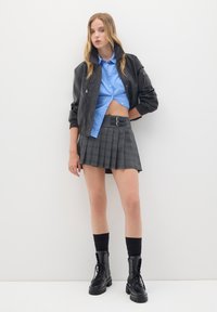 Giacca bomber nera in ecopelle, camicia croppata a righe blu, mini gonna grigia plissè con dettaglio a doppia fibbia, e stivali militari neri.