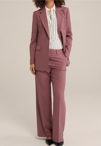 Ljust rosa skräddarsydd kostym med dubbelknäppt kavaj och vida byxor, kombinerad med en vit skjorta med svarta ränder och svarta klackar.