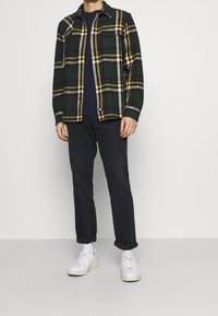 Green and yellow plaid flannel shirt with two chest pockets over a navy top, paired with black jeans and white sneakers.