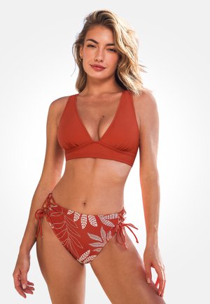 Mujer con un bikini rojo óxido, con la parte superior lisa y el pantalón estampado, de pie frente a un fondo blanco liso.