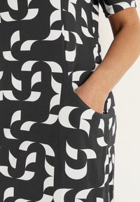 Zwarte jurk met een wit abstract geometrisch patroon, korte mouwen en zijzakken. De stof lijkt glad, lichtgewicht en gestructureerd.