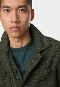 Dunkelgrüne strukturierte Jacke mit Kragen, mit einem Knopfverschluss und Fronttasche, getragen über einem blau-grünen Rundhals-Shirt.