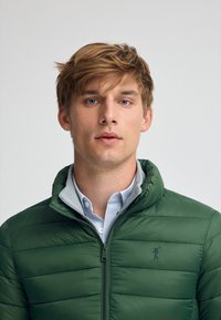 Grön puffjacka med dragkedja, som har en hög krage och ett grått innerfoder. Horisontell sömnad ger en quiltad textur.