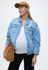 Raskaana oleva henkilö, jolla on ruskea lippalakki, vaaleansininen farkkutakki, valkoinen paita, mustat housut, ja joka pitää ruskeaa käsilaukkua, katsoo sivulle.